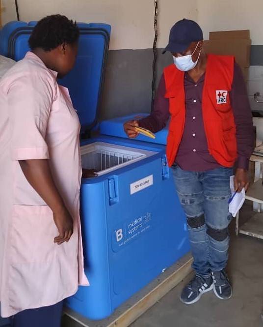 A man in a red vest, a ball cap and a mask examines a vaccine sample, standing in front of a blue refrigerated unit. A woman in a light pink lab coat stands in front of the man with her back to the camera.