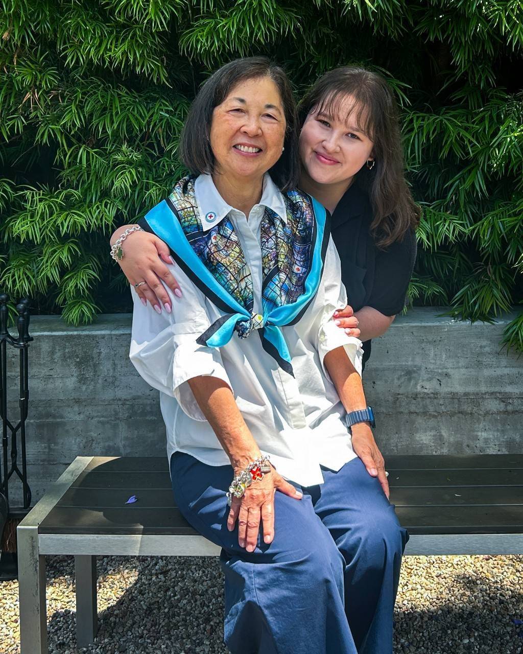 Older woman sits on bench with younger woman behind her with trees in the background.|