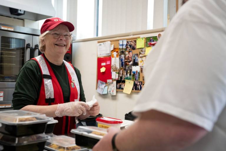 Woman in red hat and vest serves meal to man with his back to the camera.