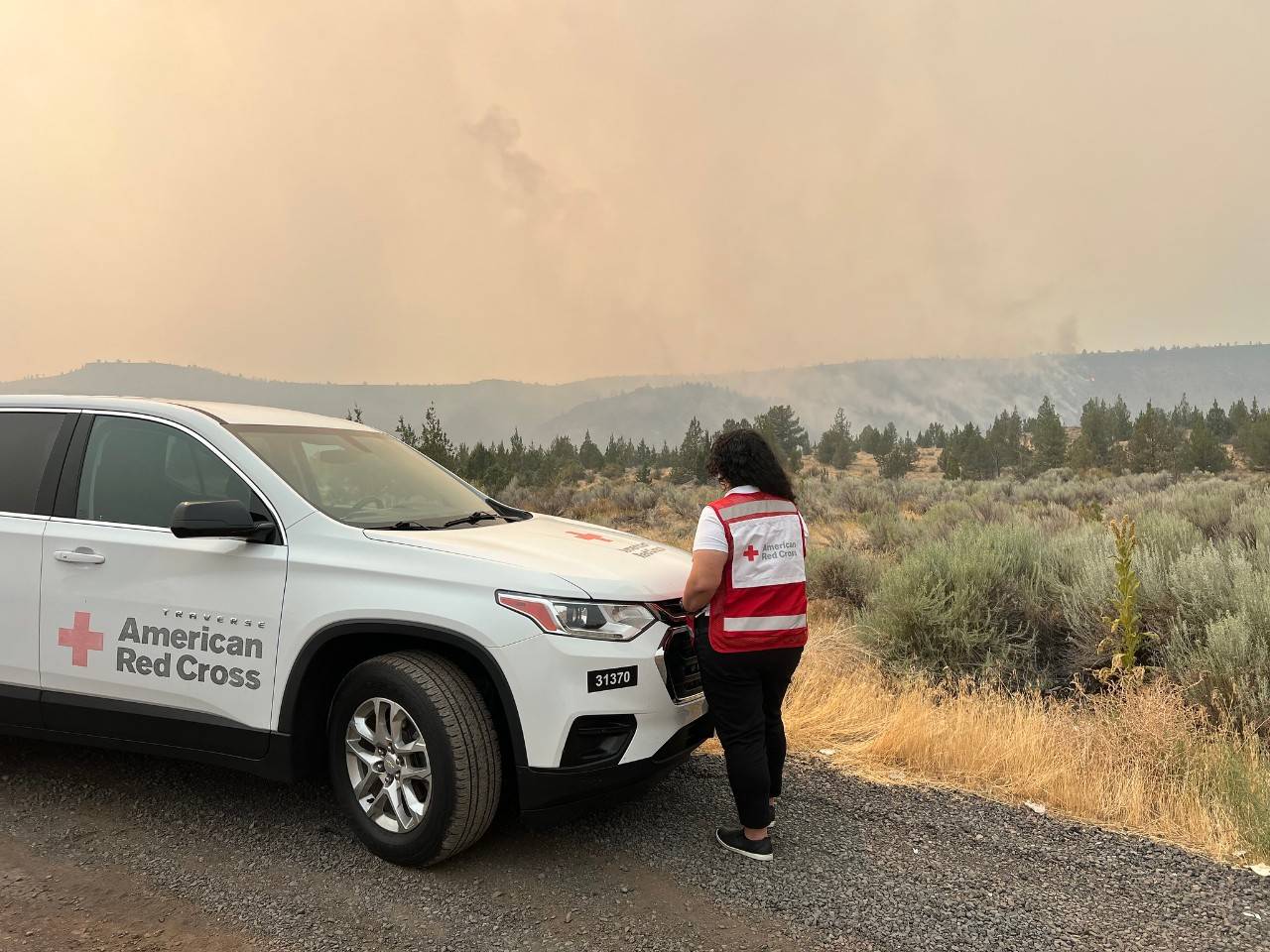 Woman in Red Cross vest stands beside a Red Cross marked vehicle.