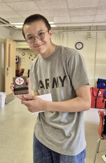 Una moneda grande de metal con una cruz roja grabada. La moneda está en un estuche pequeño de plástico; Gyan lo sostiene en alto y sonríe.