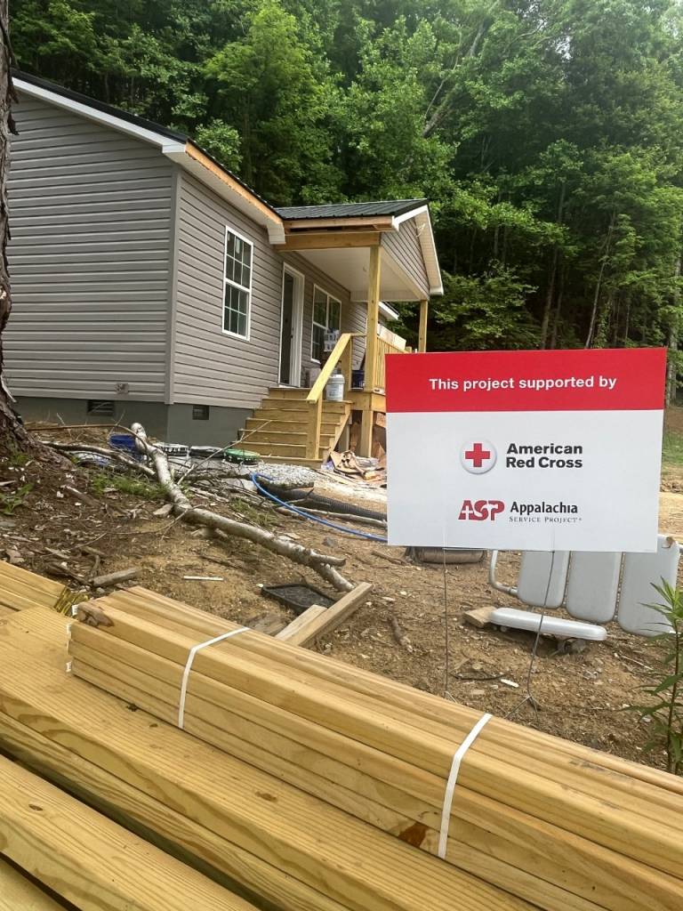 Varias pilas de madera frente a una vivienda en reparación. Un cartel rojo y blanco colocado en el jardín muestra el texto «Este proyecto cuenta con el apoyo de» y los logotipos de la Cruz Roja Americana y Appalachia Service Project, que colaboran en los trabajos de reparación.