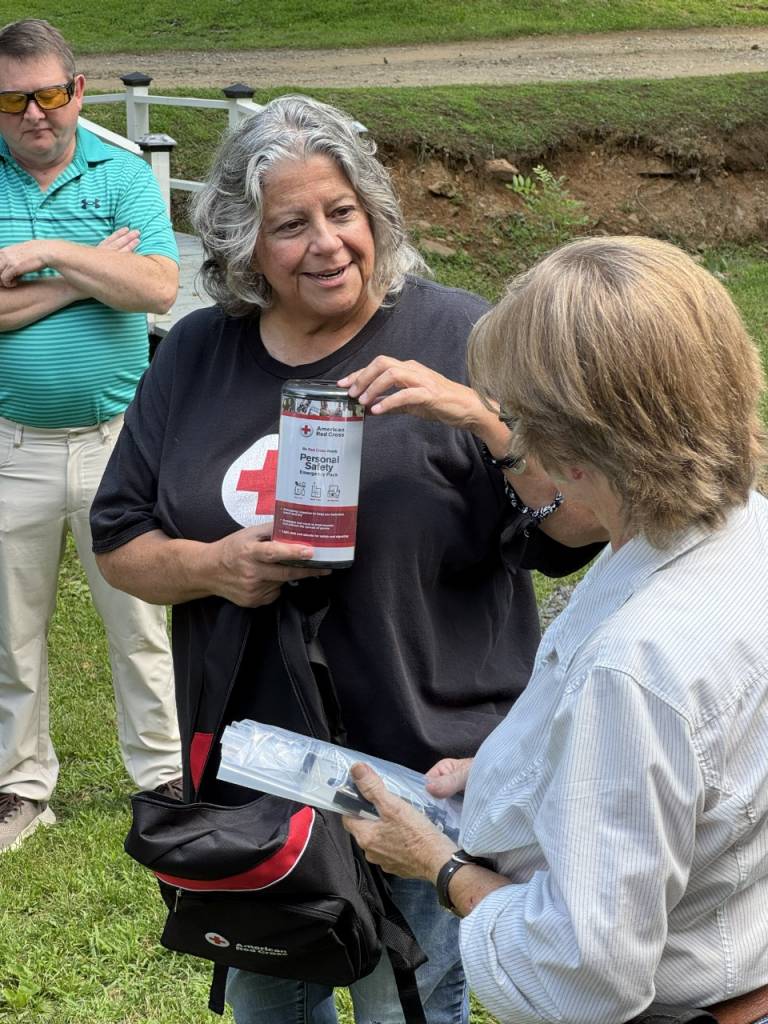 Una mujer de cabello canoso y camiseta de la Cruz Roja sosteniendo un kit de seguridad personal mientras comparte información sobre preparación ante emergencias con otra mujer de pelo corto y camisa blanca de botones.