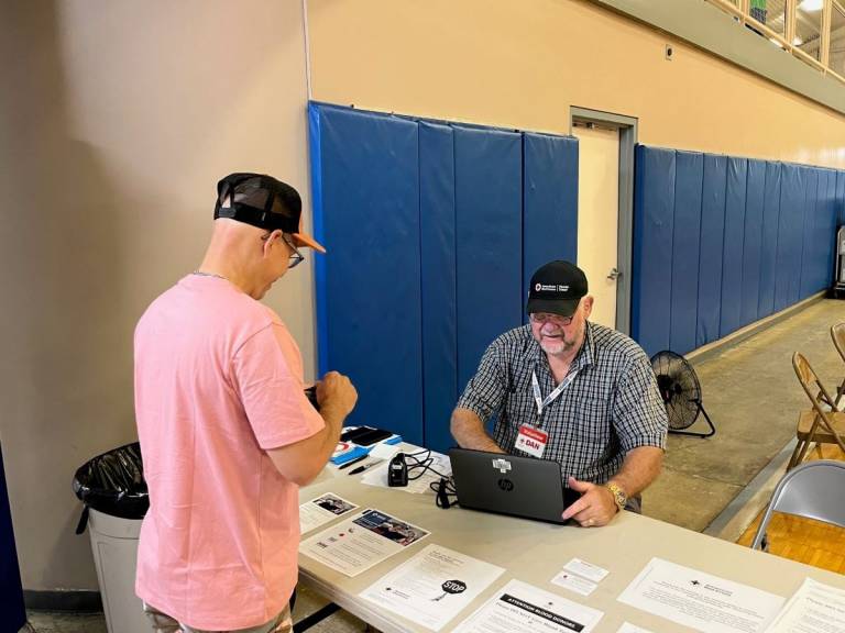 man in Red Cross badge sits at table while man in pink shirt stands