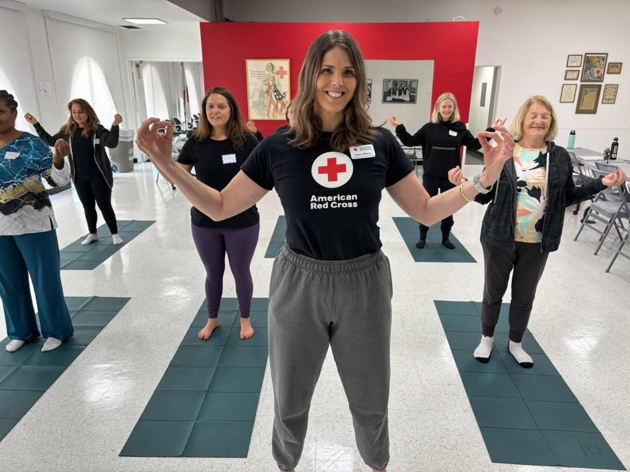 Future Mindful Movement workshop instructors stand on yoga mats with their arms outstretched in an open classroom. Participants are in training to instruct Red Cross Mindful Movement program workshops.