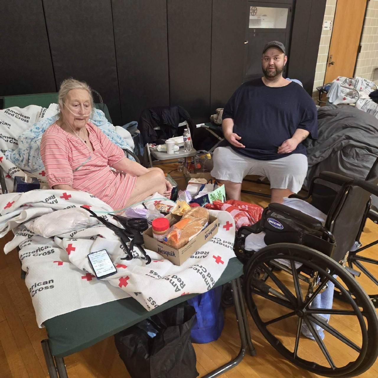 A man sits across from a woman sitting on bed covered with Red Cross blankets.
