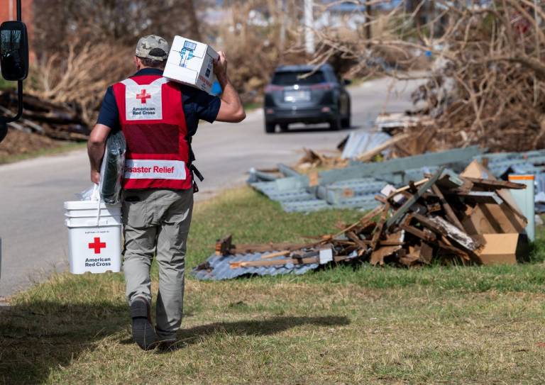 Man in Red Cross vest carries two clean-up kits through a neighborhood with storm damage