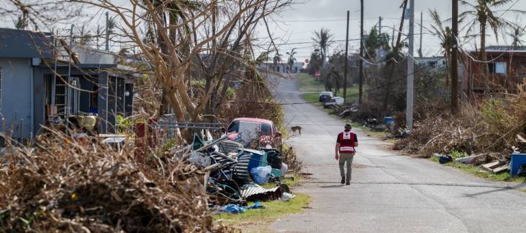A man in a Red Cross vest walks down a street full of damage from the storm.