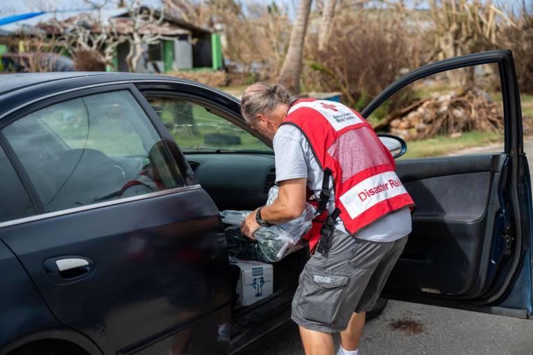 A man in a Red Cross vest loads relief supplies into the front passenger seat of a car.