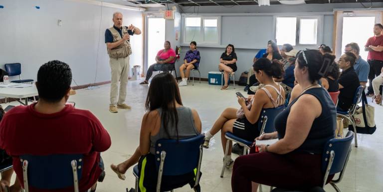 A group of people sit and listen to a man in a Red Cross vest.