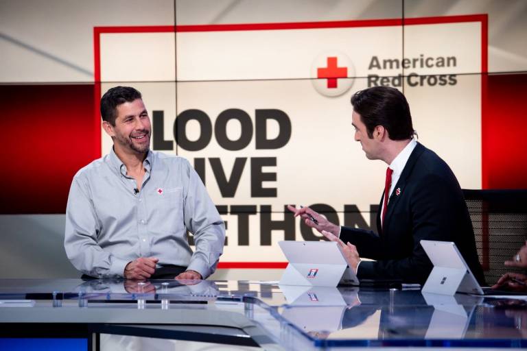 man in Red Cross shirt speaks to news anchor