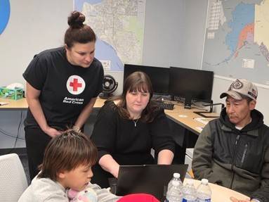 four people, one in a Red Cross tee shirt, gather around a laptop sitting on a desk.
