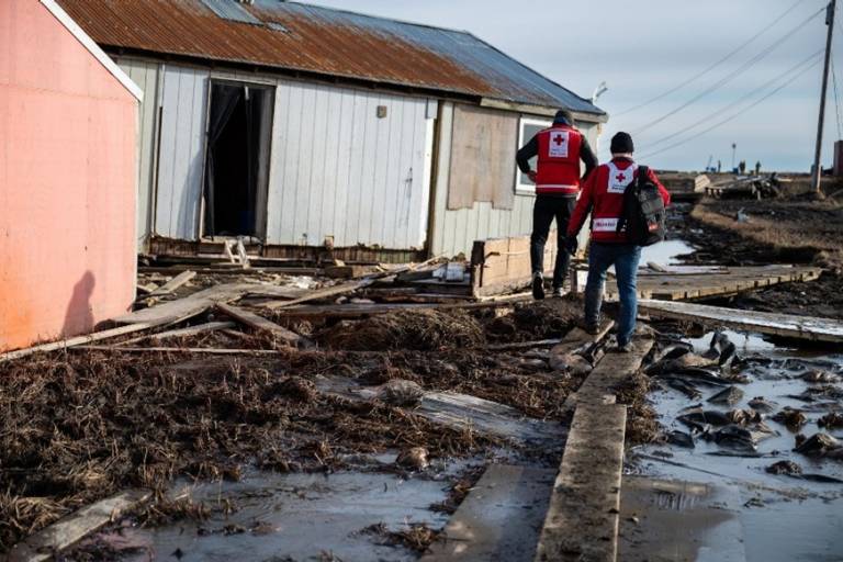Two men in Red Cross vests walk down an area filled with rubble from flooding.