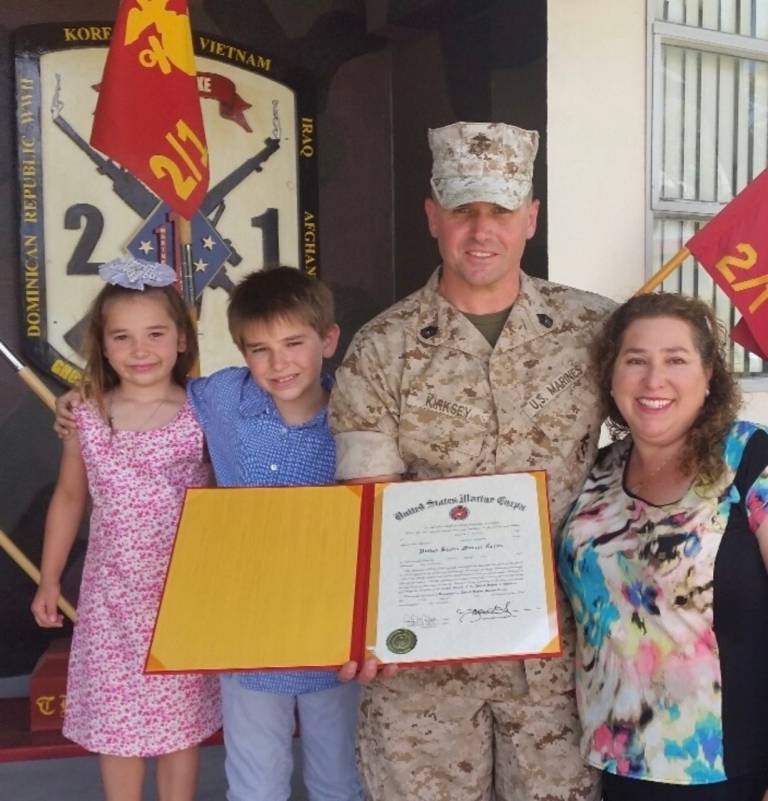 a young Hannah in a family photo with her dad (in uniform), mom and brother. They have their arms around each other and smile. Their dad holds a service award certificate.