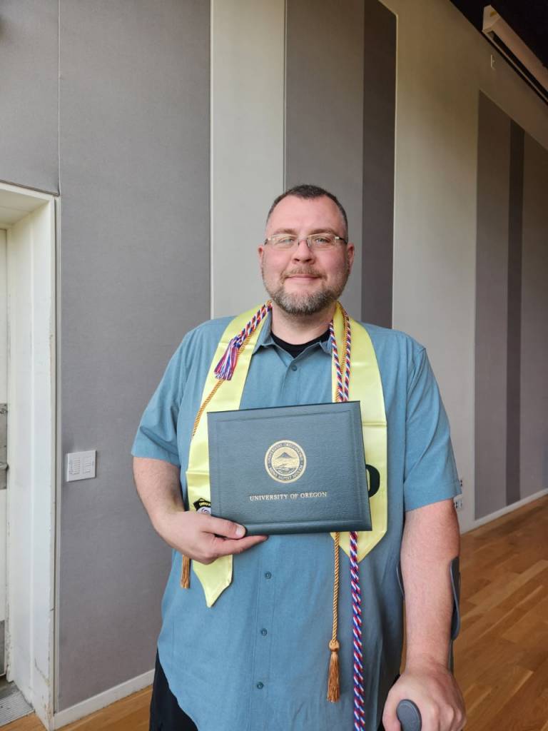 Spc. Rose more than 20 years later. He still wears glasses but he has a buzzcut and trimmed beard. In one hand he supports his weight with a cane, the other holds his diploma from University of Oregon. 