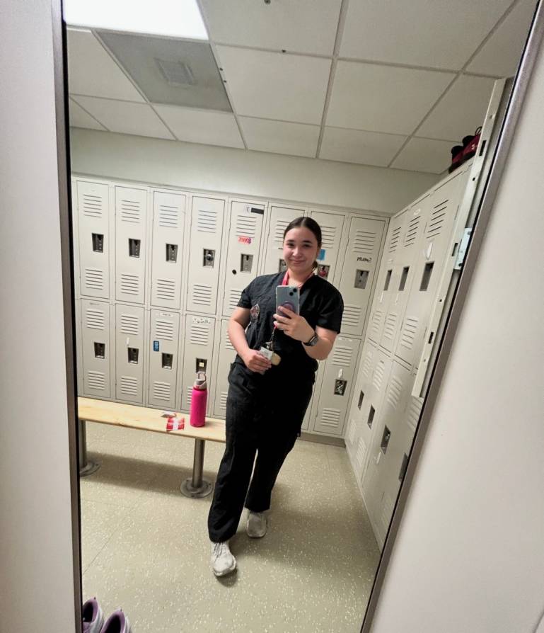 Hannah takes a selfie in the mirror of a locker room. She has her hair pulled back and is in scrubs.