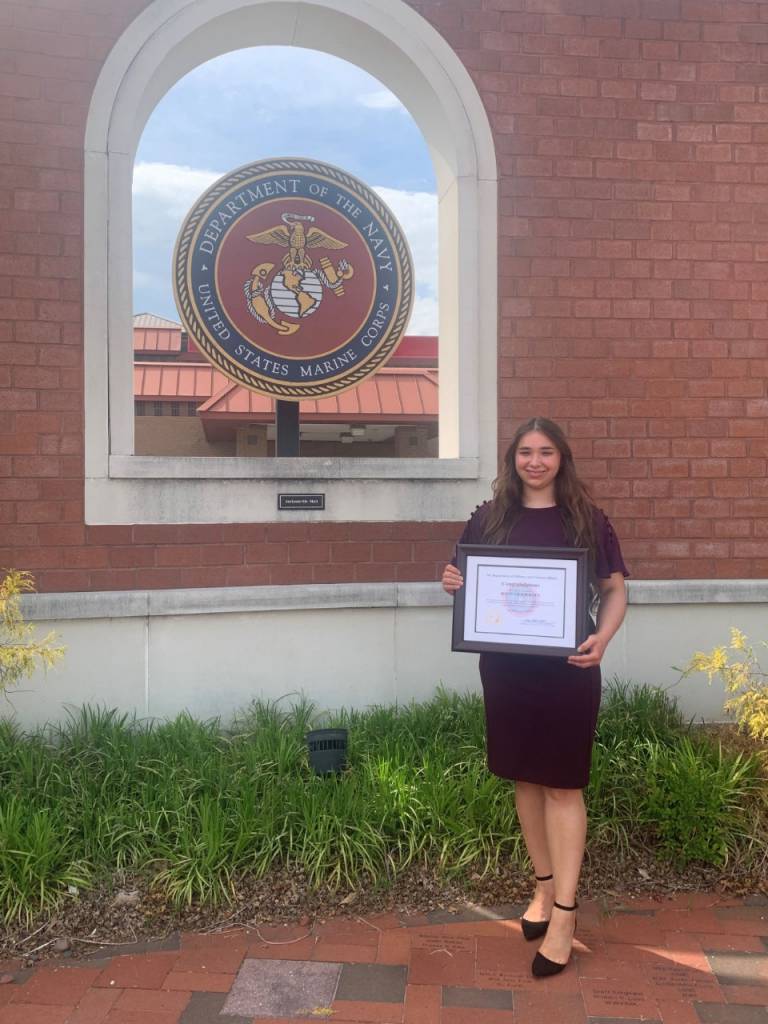 Hannah in formal clothes holds an award certificate, standing beside the seal of the U.S. Marine Corps.