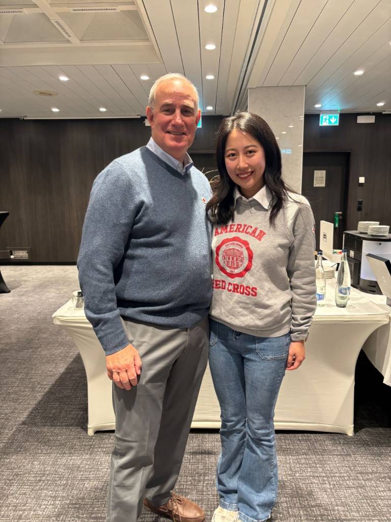 A young woman in a Red Cross sweatshirt stands with a man in a gray sweater