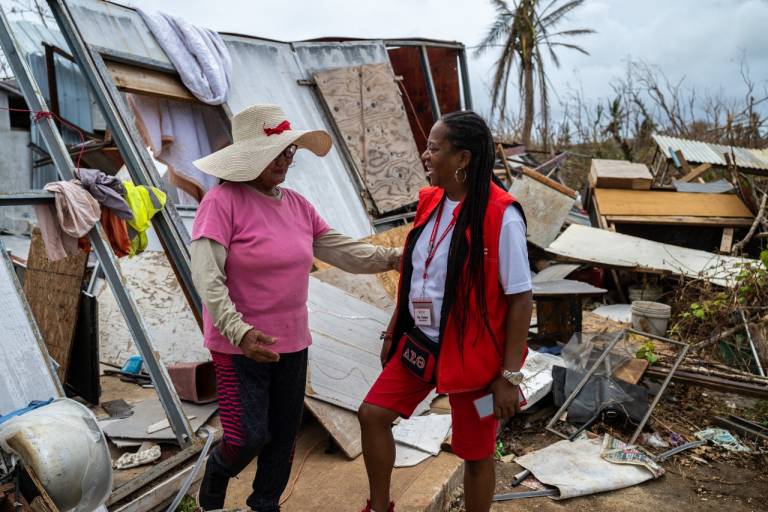 A Red Cross volunteer wearing all red talks with a woman in front of a damaged home.