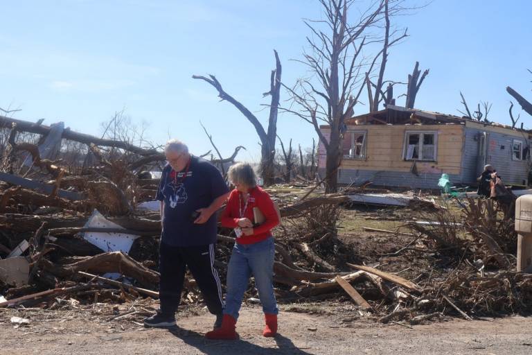 un hombre y una mujer con chaleco de la Cruz Roja Americana caminan junto a los daños causados por la tormenta.