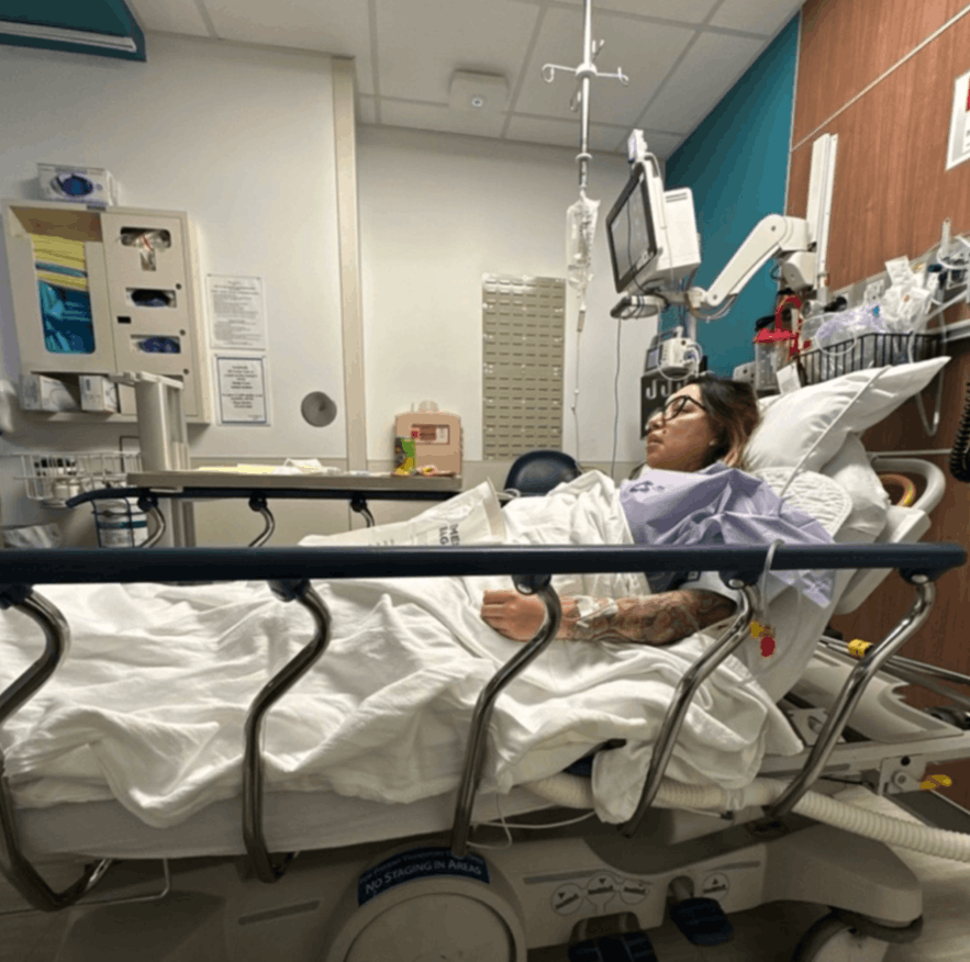 A woman lying in a hospital bed, recovering from surgery. Her left arm is connected to an IV bag. There is a monitor at her bedside, along with other medical equipment.
