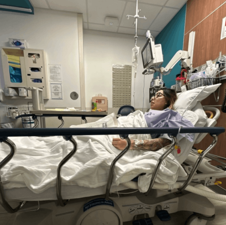 A woman lying in a hospital bed, recovering from surgery. Her left arm is connected to an IV bag. There is a monitor at her bedside, along with other medical equipment.