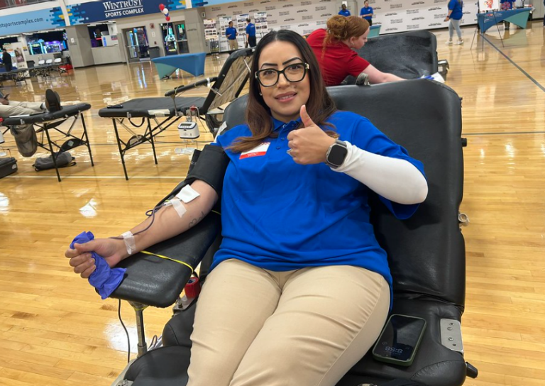 A woman sitting in a black chair at a blood donation center. One of her arms is connected to a blood collection bag, and she is making an “OK” sign with her other hand. In the background of the gym serving as the donation center, several phlebotomists can be seen, along with part of a person lying on a donation stretcher.