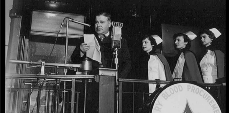 Fotografía en blanco y negro de un hombre con traje y mujeres con capas de la Cruz Roja a bordo de la parte trasera de un tren.