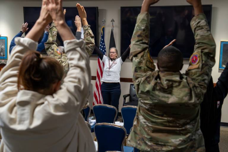A Red Cross volunteer leads a group of uniformed service members and civilians through a movement in the Mindful Movement workshop. The group has their arms stretched above their heads.