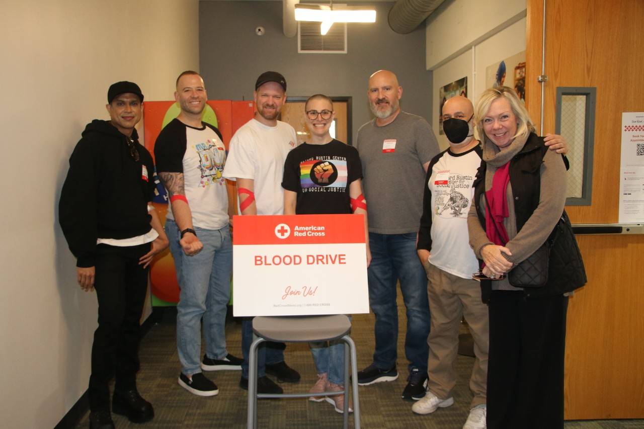 A group of seven smiling blood donors stand around a blood drive sign showing their blood donation arm bandages. 