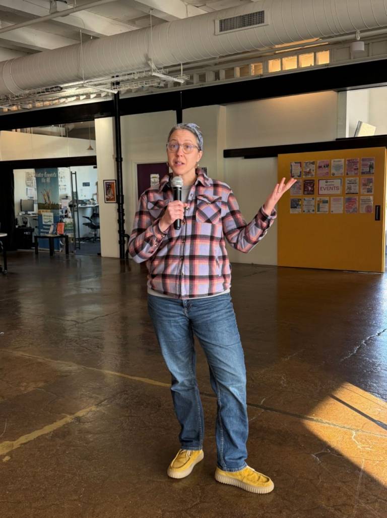 Hilary Otey in a large meeting room at Queermunity community center speaking into a microphone presenting at a community event. 