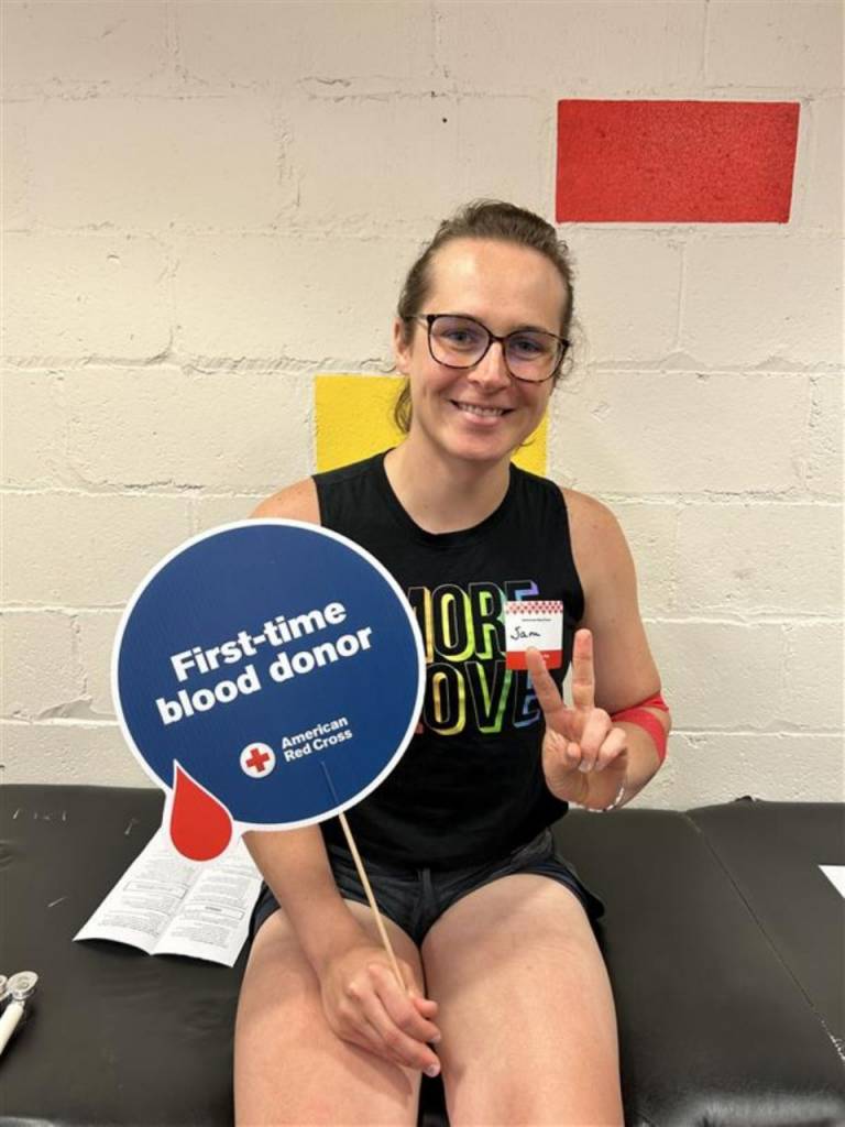 Sam sits on a donor bed after donation, smiling and holding a sign that reads “First time blood donor.”