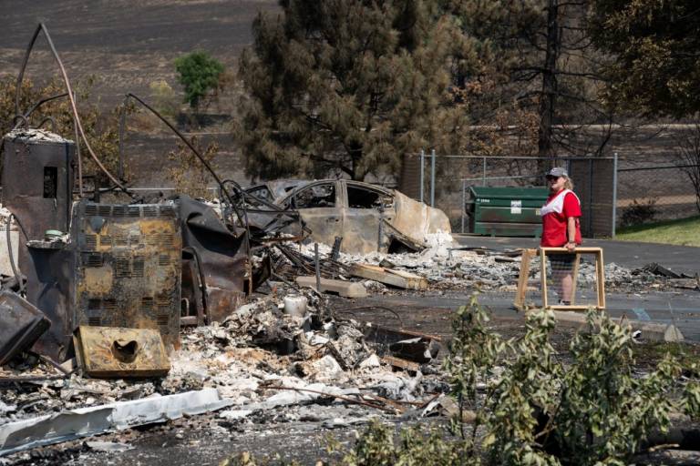 Woman in Red Cross vest stands near the charred remains of a home destroyed by a wildfire.