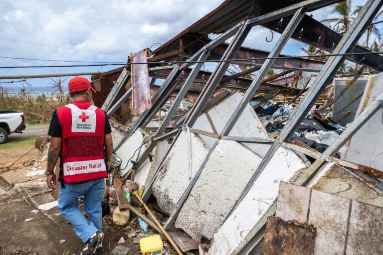 Man in Red Cross vest walks past a damaged structure on Tinian.