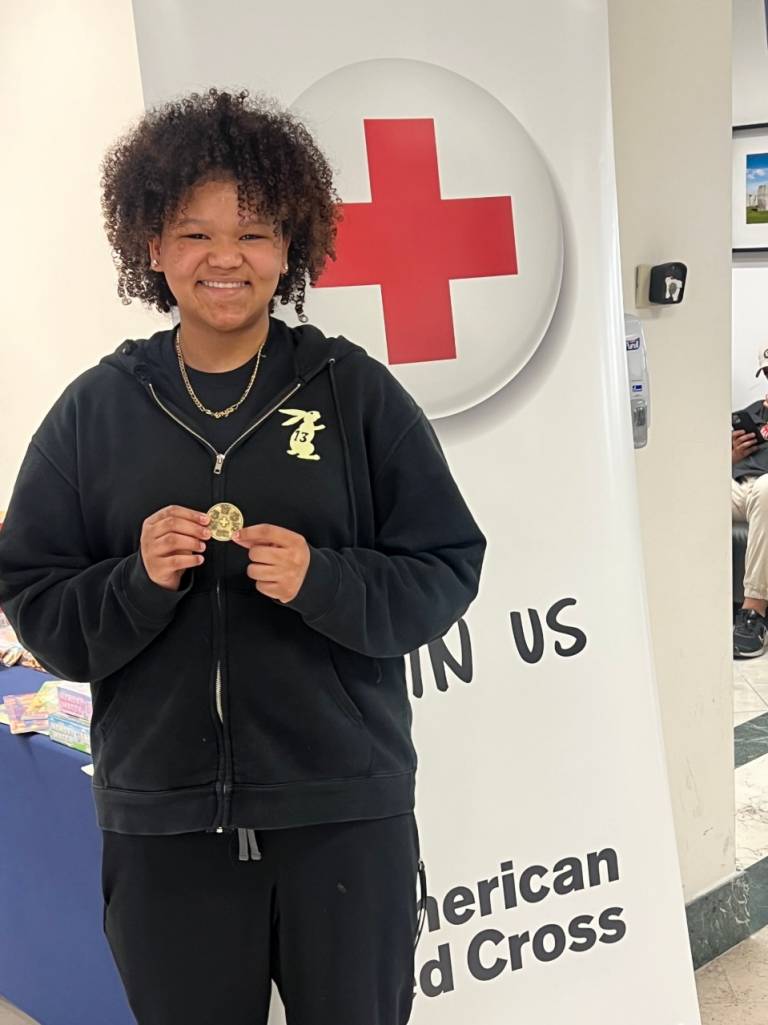 A teen stands in front of a Red Cross banner. The teen holds a Red Cross challenge coin in her hands.