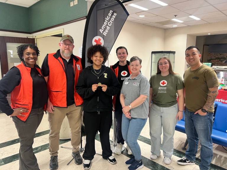 A group of people, some in Red Cross vests, stand with a teen in front of a Red Cross banner. The teen holds a Red Cross challenge coin in her hands. 