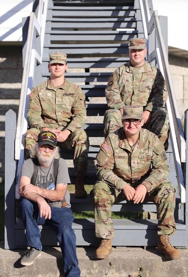 Four men sit on stairs together smiling. Three are in military uniforms and one is wearing a Vietnam Veteran hat.