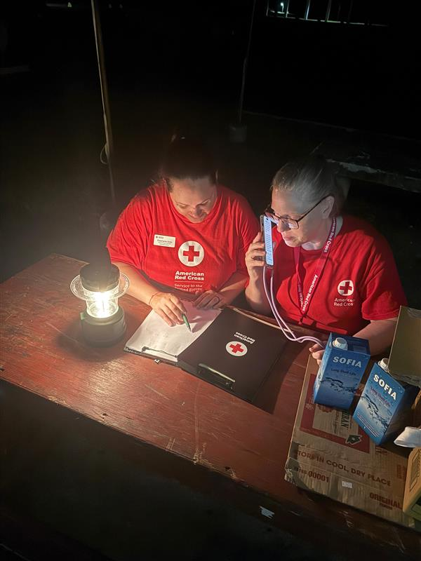 Two SAF Red Crossers sitting in the dark, huddle around a lamp while the power is out. One is on the phone while the other takes notes. 