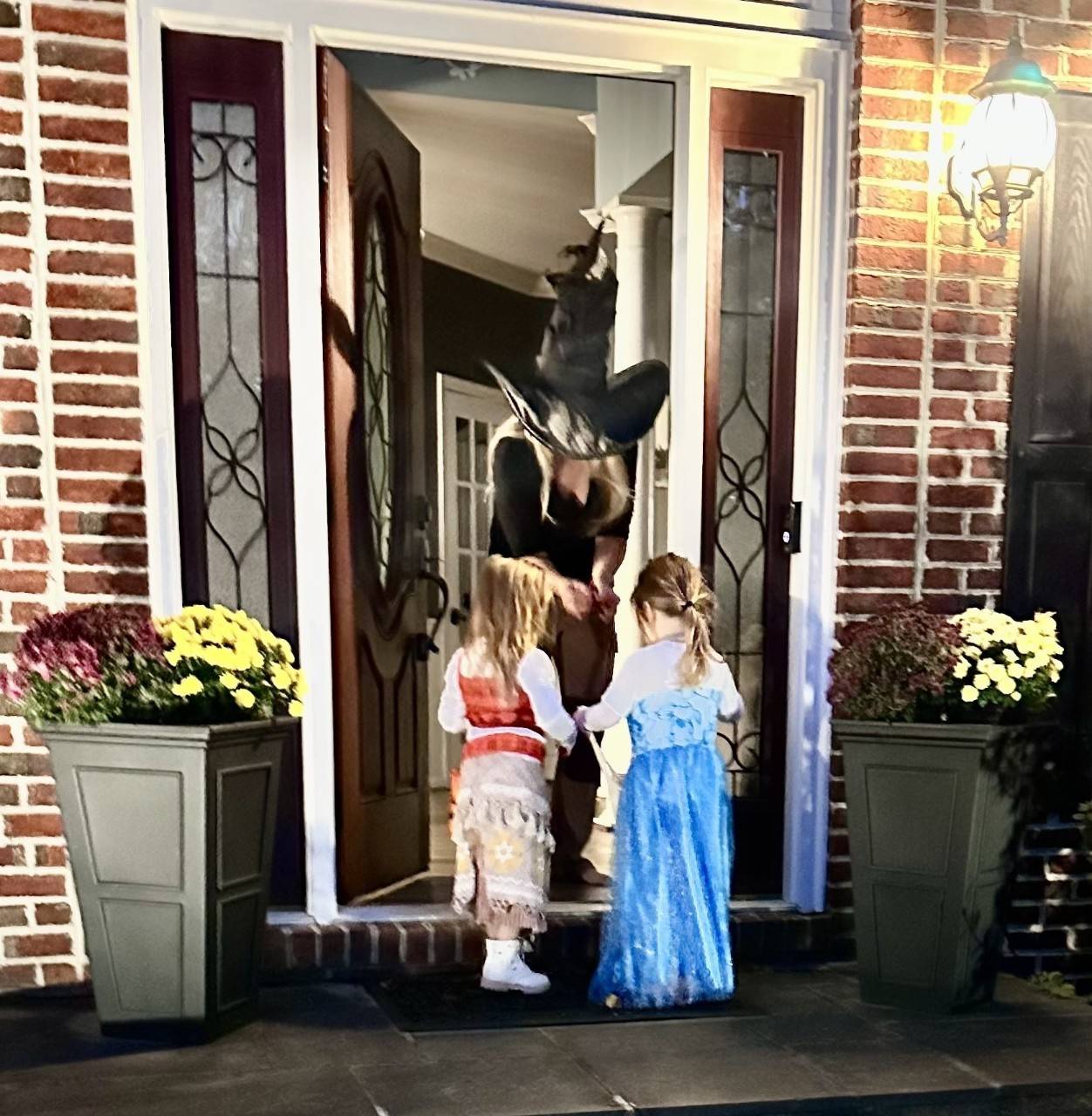 Two children stand at the doorstep of a brick home in Halloween costumes. One is wearing a princess dress. An adult at the door wearing a witch costume drops candy in a trick-or-treat bag.