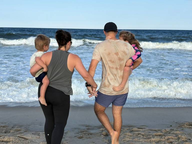 Two adults holding hands and carrying two children stand by the shoreline of a beach. Their backs face the camera. 