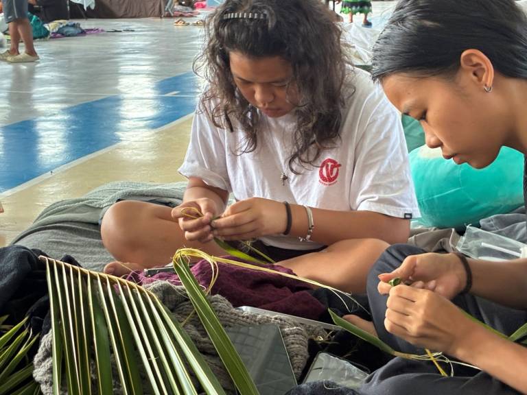 Two girls sit with pile of palm leaves which they weave into flowers