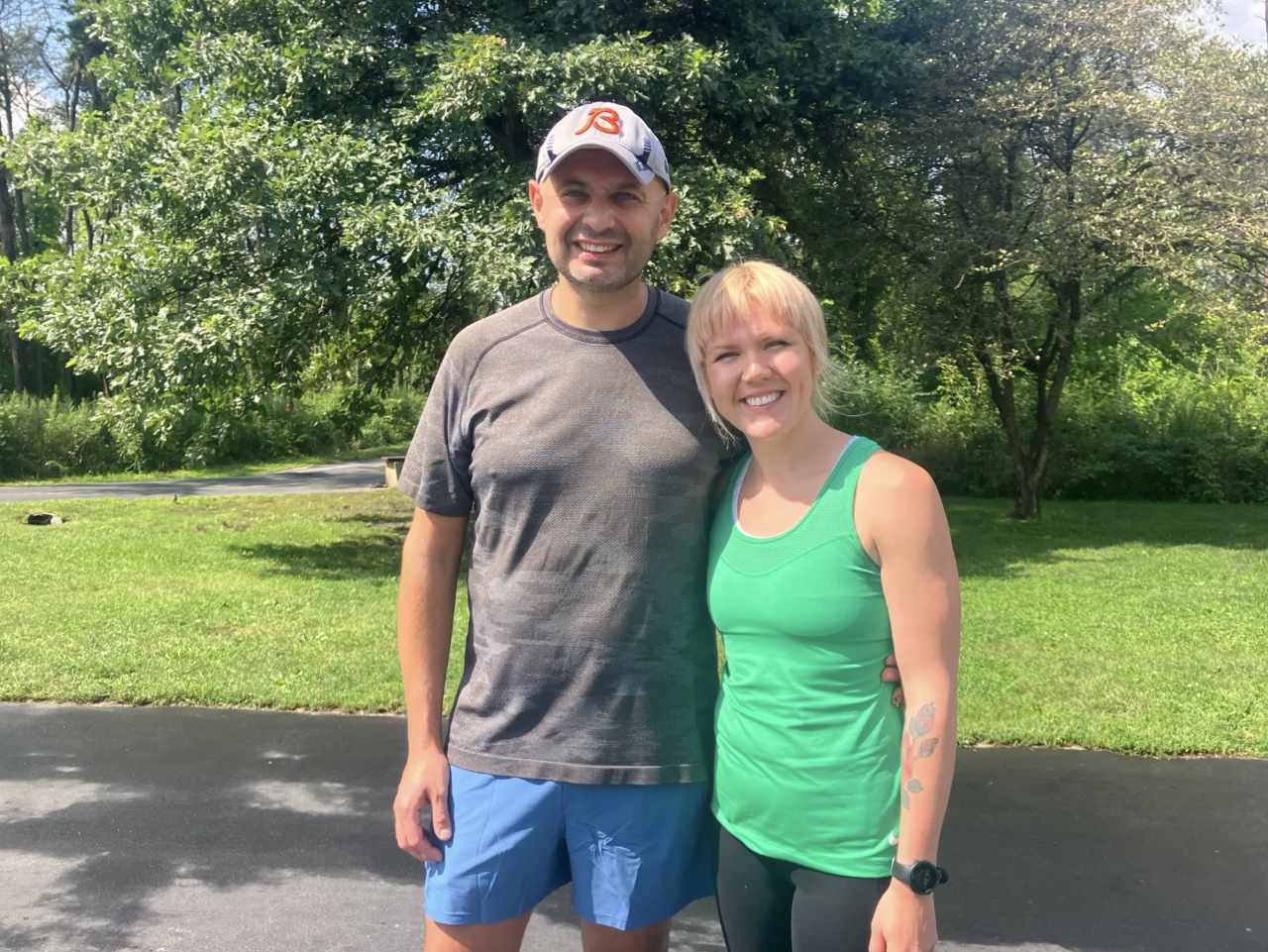 Two people stand smiling on an outdoor trail with greenery and trees behind them. One is wearing a baseball cap, the other is wearing a green tank top.