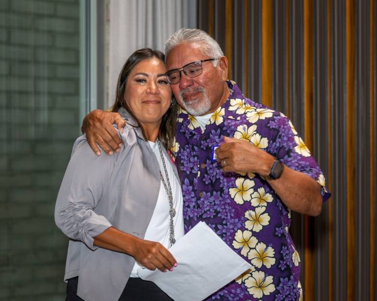 Two people embrace in a tearful, emotional moment on a stage. One wears a floral shirt, the other is holding a certificate.