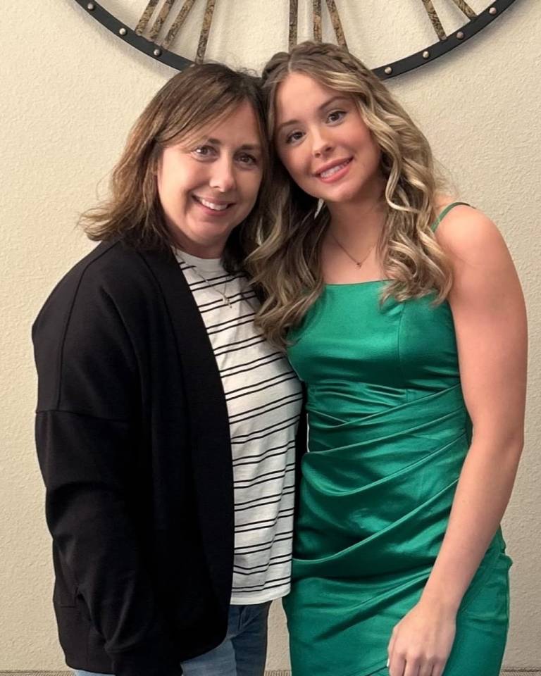 Two people stand inside a room with a wall behind them with a giant clock on it. One person is in a green formal dress.