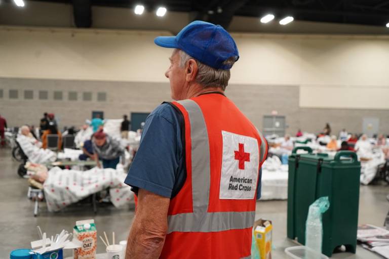 Man in Red Cross vest looks out on people in disaster shelter