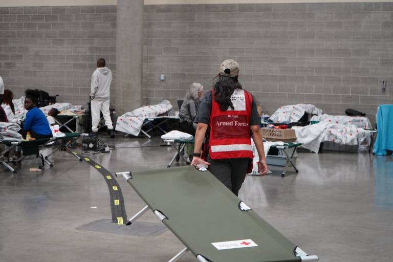 Red Crosser in a disaster vest sets up cots in a shelter.