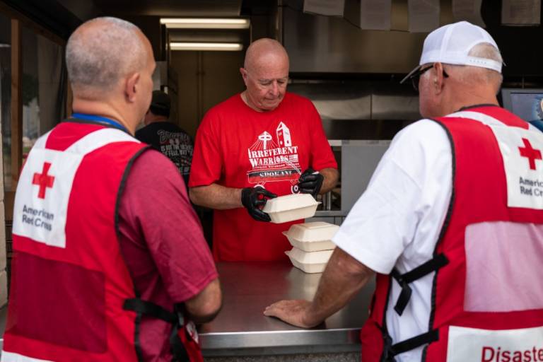Two men in Red Cross shirts get meals to deliver to wildfire victims from a man in a VFW shirt.