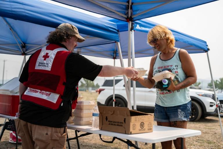 Man in Red Cross vest hands meals to man with blond hair.