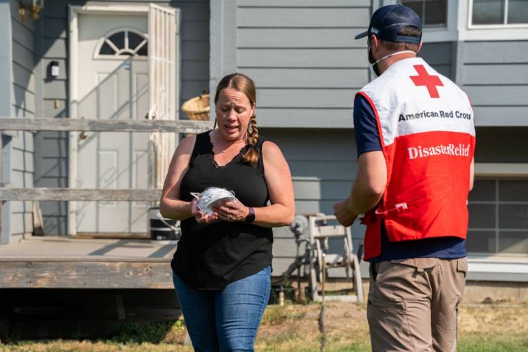 Man in Red Cross vest hands mask to woman in black shirt.