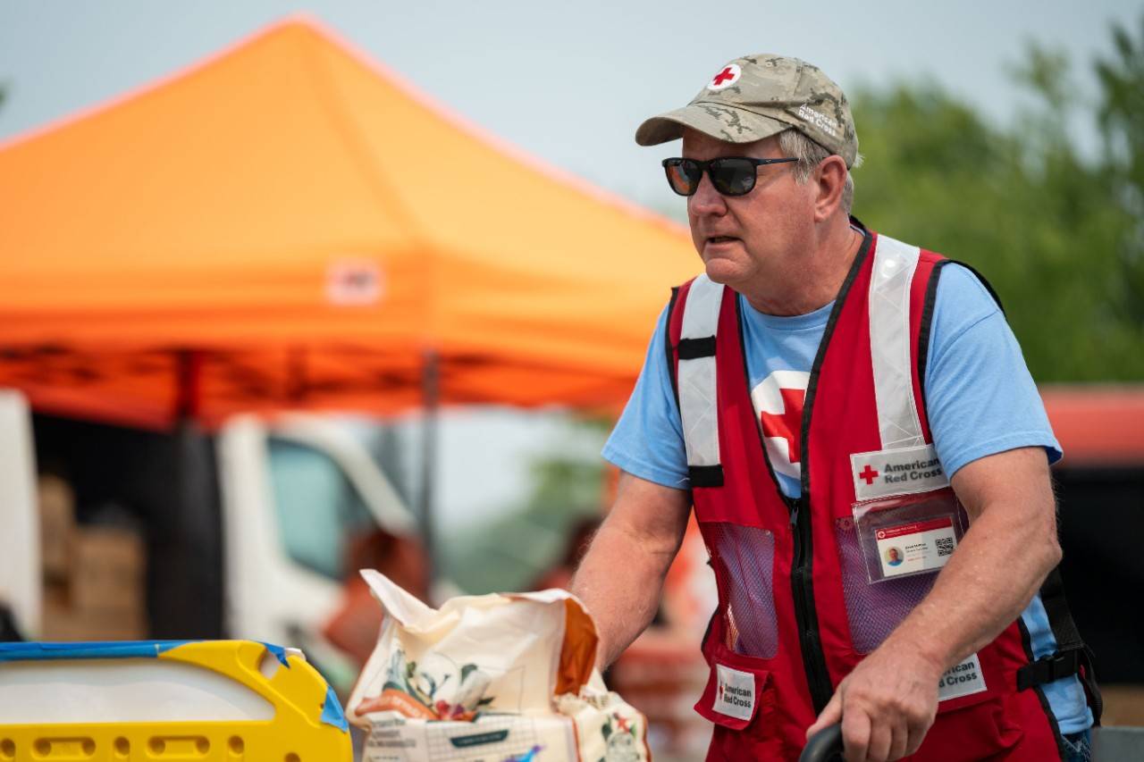 Man in Red Cross vest pushes dolly filled with pet supplies.